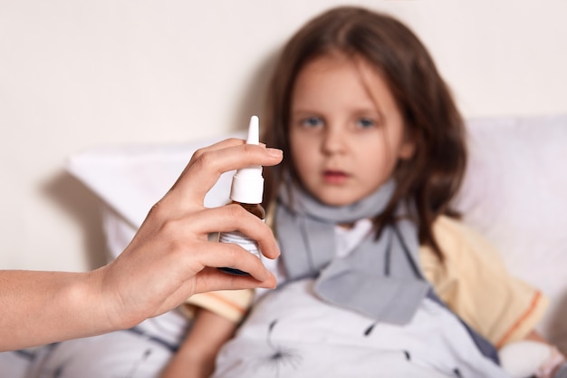 Little girl lying in bed, her mom treating her runny nose with nasal spray, dark haired female child looking at camera