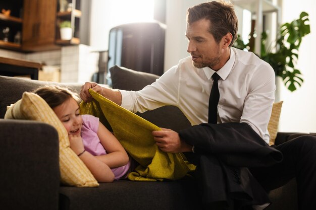 Little girl lying down on the sofa while her father is covering her with a blanket before going to work Focus is on man