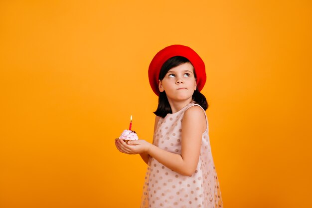 Little girl making birthday wish.  brunette kid holding cake with candle on yellow wall.