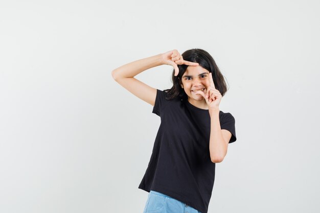 Little girl making frame gesture in black t-shirt, shorts and looking cheerful , front view.