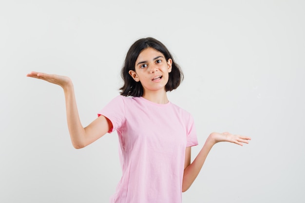 Little girl making scales gesture in pink t-shirt and looking confused. front view.