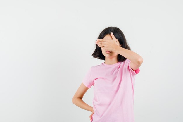 Little girl in pink t-shirt holding hand on eyes and looking scared , front view.