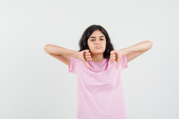 ピンクのTシャツを着た少女は、二重の親指を下に向けて、がっかりしているように見えます。正面図。