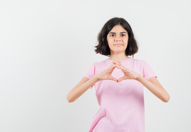 Little girl in pink t-shirt showing heart gesture and looking cheery , front view.