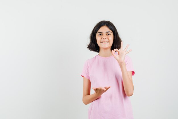 Маленькая девочка в розовой футболке показывает знак ОК, держит ладонь открытой и выглядит веселой, вид спереди.