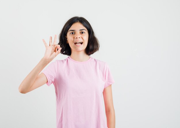 Little girl in pink t-shirt showing ok sign and looking cheery , front view.