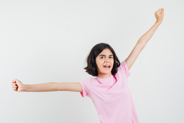 Маленькая девочка в розовой футболке показывает жест успеха, протягивая руки и выглядит счастливым, вид спереди.