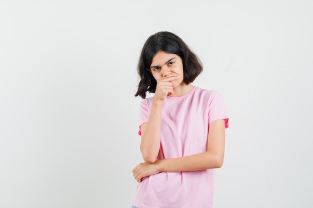 Маленькая девочка в розовой футболке стоя в позе мышления и выглядела обеспокоенной, вид спереди.