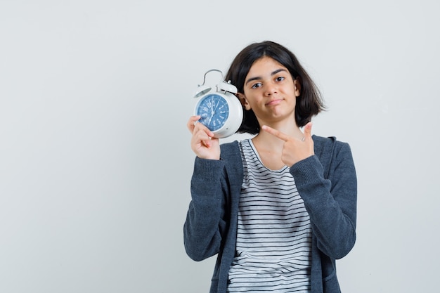 Little girl pointing at alarm clock in t-shirt, jacket and looking confident ,