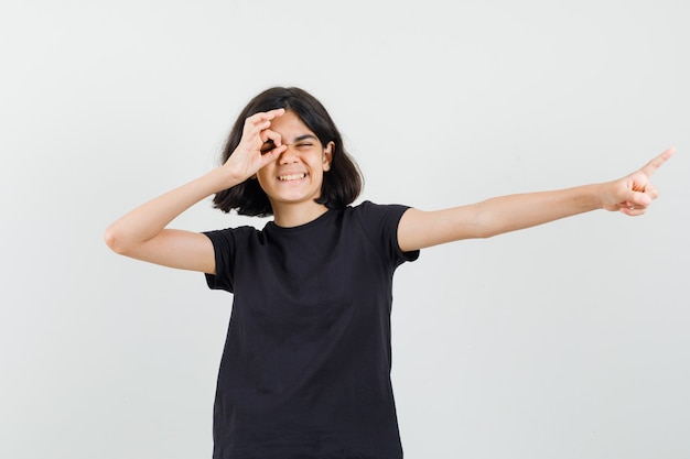 Little girl pointing aside, showing ok sign on eye in black t-shirt , front view.