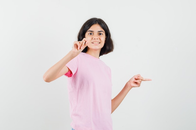 Little girl pointing aside, showing small size sign in pink t-shirt and looking cheery , front view.