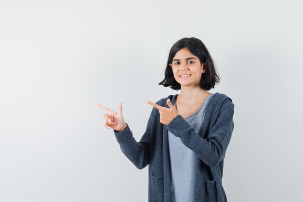 Little girl pointing aside in t-shirt, jacket and looking cheerful.