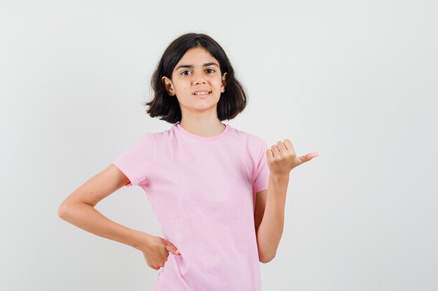 Маленькая девочка указывая в сторону большим пальцем в розовой футболке и выглядит уверенно, вид спереди.