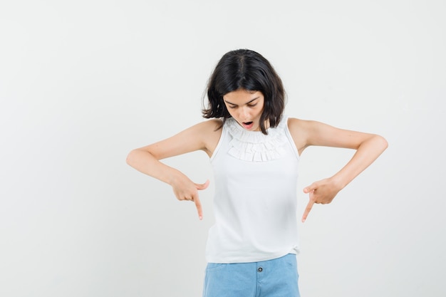 Little girl pointing down in white blouse, shorts and looking surprised , front view.