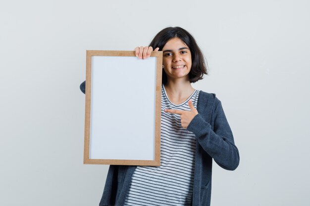 Little girl pointing at empty frame in t-shirt, jacket and looking glad ,