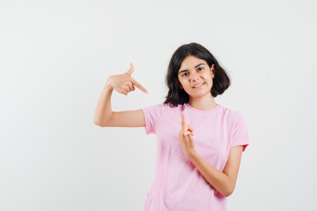 Little girl pointing fingers up and down in pink t-shirt and looking optimistic , front view.