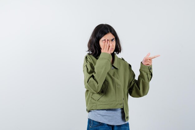 Little girl pointing to the right side, covering eye with hand in coat, t-shirt, jeans and looking confident , front view.