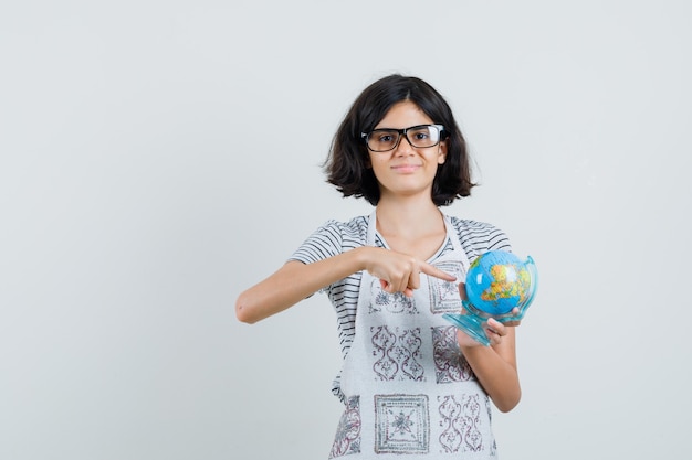 Little girl pointing at school globe in t-shirt, apron and looking confident.