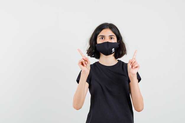 Little girl pointing up in black t-shirt, mask and looking sensible. front view.