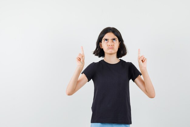 Little girl pointing up in black t-shirt, shorts and looking gloomy , front view.