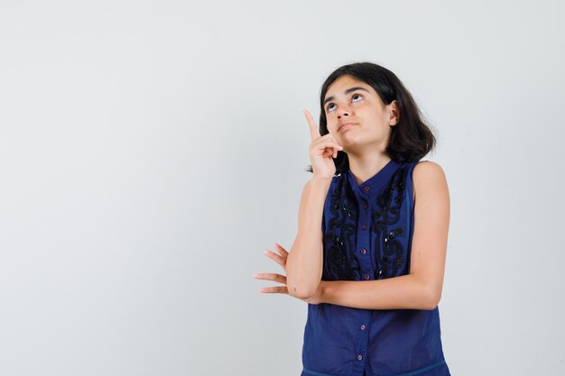 Little girl pointing up in blue blouse and looking curious.