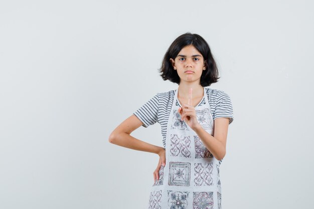 Little girl pointing up in t-shirt, apron and looking serious