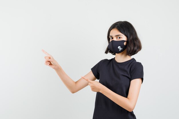 Little girl pointing at upper left corner in black t-shirt, mask and looking confident. front view.