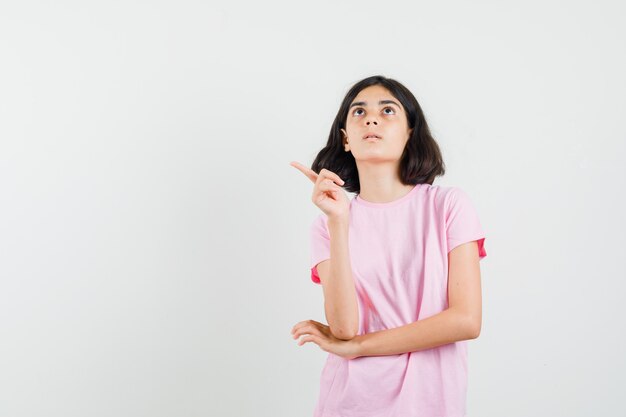 Little girl pointing at upper left corner in pink t-shirt and looking focused. front view.