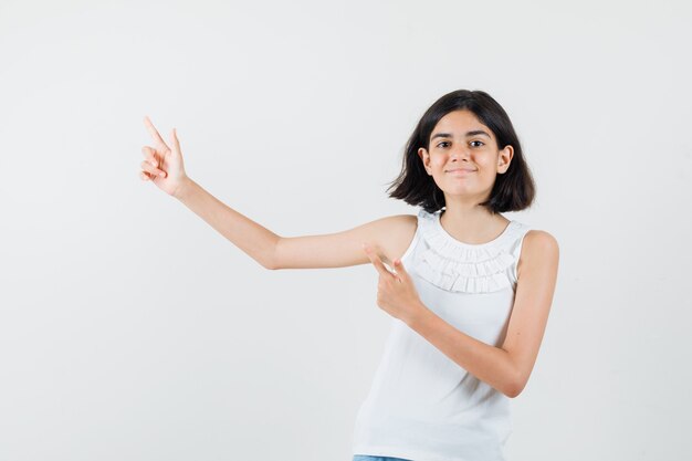 Little girl pointing at upper left corner in white blouse and looking cheery , front view.