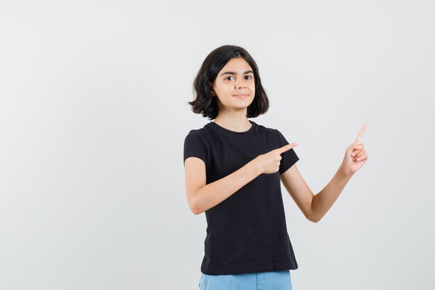 Маленькая девочка, указывая на верхний правый угол в черной футболке, шортах и уверенно, вид спереди.