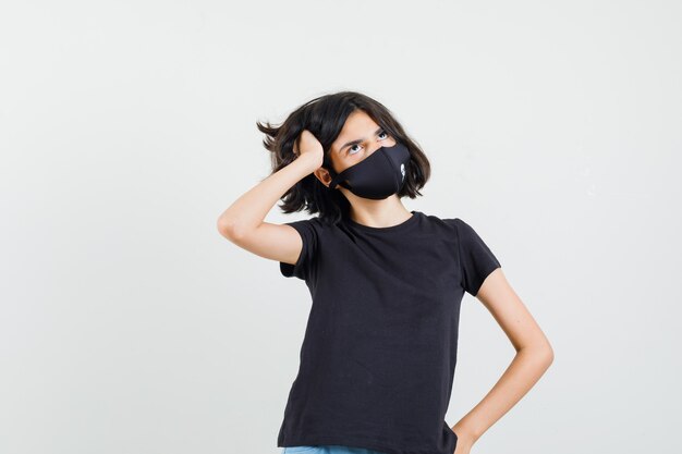 Little girl posing while standing in black t-shirt, mask and looking pensive , front view.