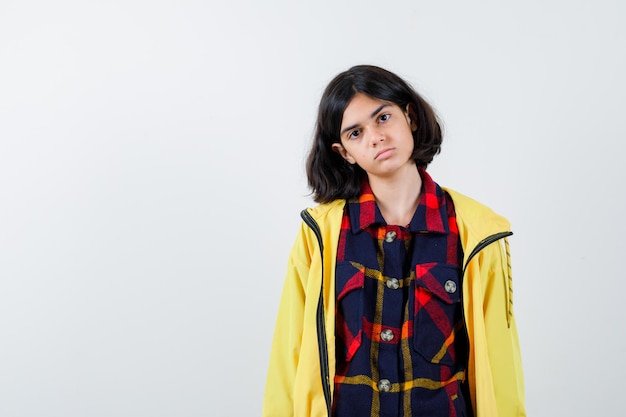 Little girl posing while standing in checked shirt, jacket and looking wistful , front view.