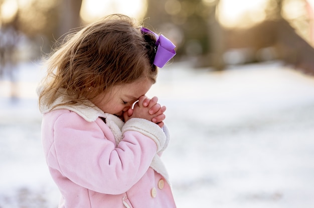 ぼやけた距離で日光の下で雪に覆われた庭で祈る少女