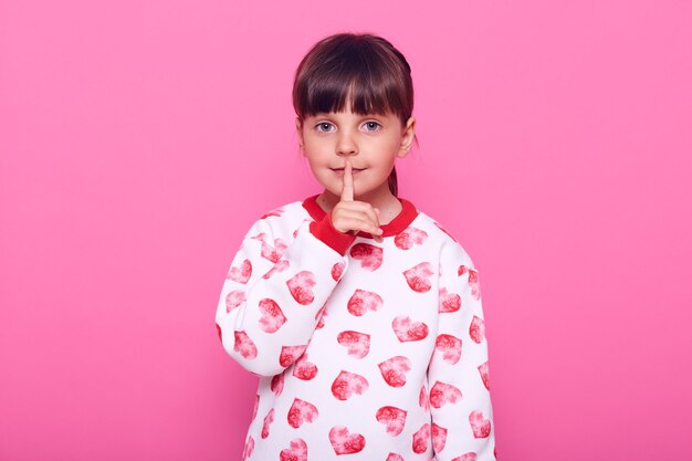 Little girl of preschool age, puts her finger near her lips, dresses in sweater, isolated over pink wall.