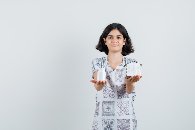 Little girl presenting house model, bottle of pills in t-shirt, apron and looking gentle