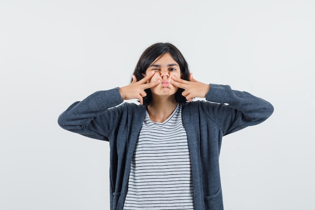 Little girl pressing fingers on cheeks in t-shirt, jacket and looking pensive