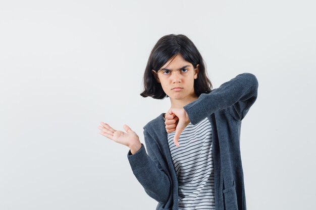 何かを持っているふりをして、Tシャツ、ジャケットで親指を下に見せて、真剣に見える少女