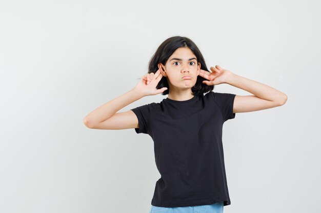 Little girl pushing ears with fingers in black t-shirt, shorts and looking funny , front view.