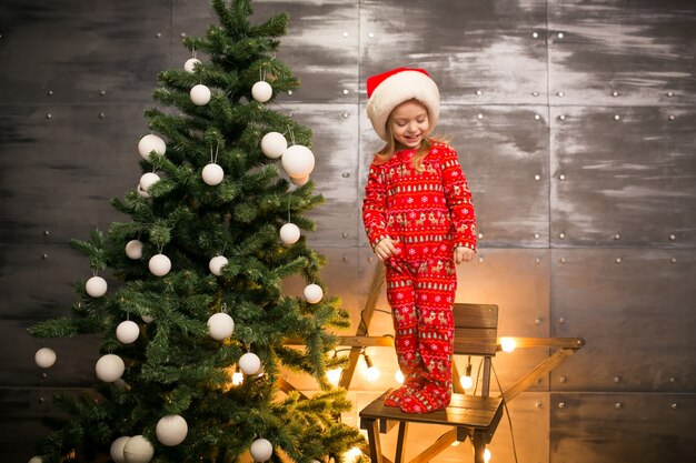 Little girl in pyjamas by the Christmas tree on a wooden chair