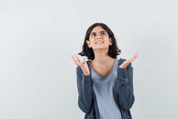 Little girl raising hands while looking up in t-shirt, jacket and looking grateful