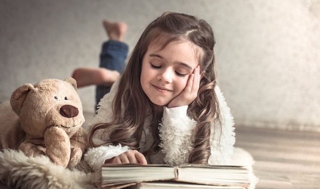 Bambina che legge un libro con un orsacchiotto sul pavimento, concetto di relax e amicizia