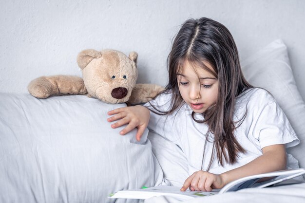 小さな女の子は朝、ベッドでテディベアと一緒に本を読みます