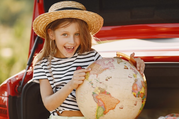 無料写真 休暇に行く準備ができている少女。赤い車の子供。地球儀と帽子を持つ少女。