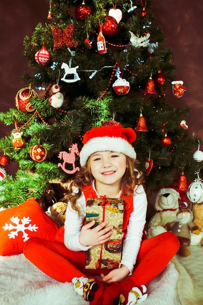 La bambina in vestito rosso si siede con una scatola attuale prima di un albero di natale