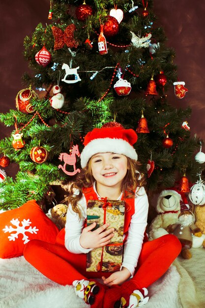 Little girl in red dress sits with a present box before a Christmas tree 