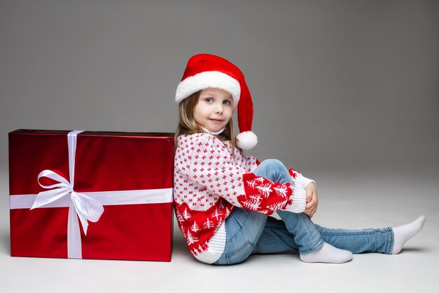 Little girl in Santa hat and sweater with winter pattern leaning on red Christmas present with white bow. Studio shot on grey wall