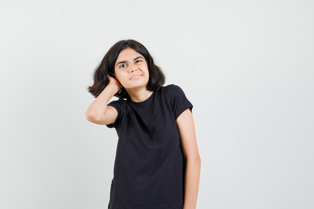 Little girl scratching head in black t-shirt and looking pensive. front view.