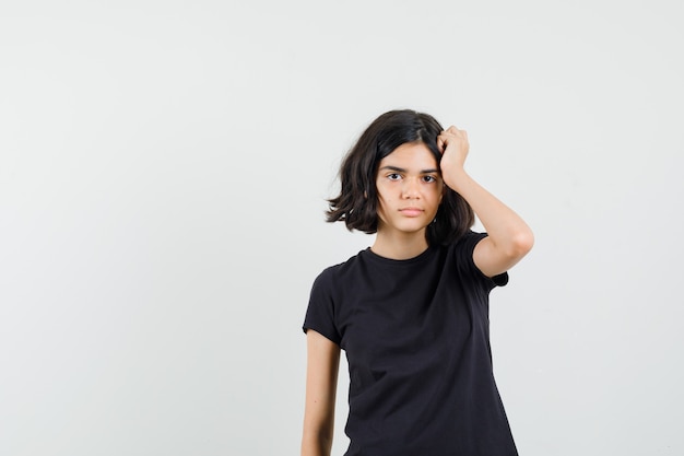 Little girl scratching head in black t-shirt and looking pensive , front view.