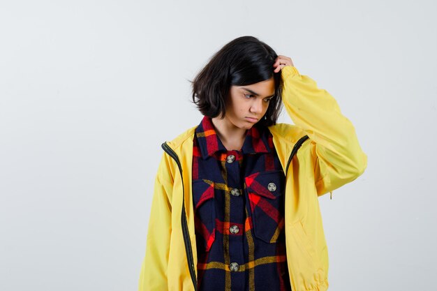 Little girl scratching head in checked shirt, jacket and looking thoughtful , front view.