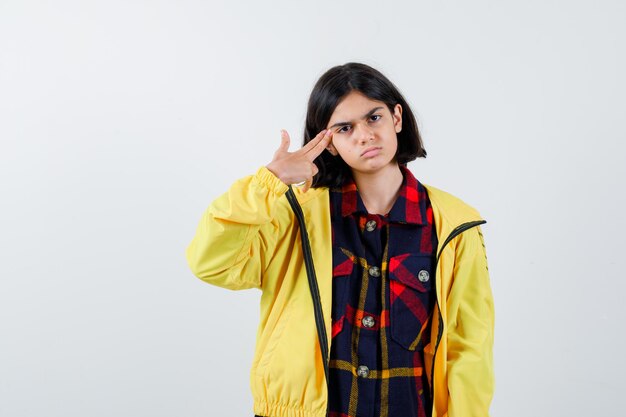 Little girl shooting herself with hand gun in checked shirt, jacket and looking hopeless , front view.